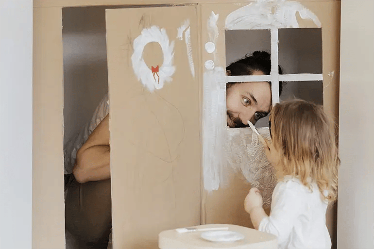a father playing with his daughter while painting a cardboard box house and the father's nose