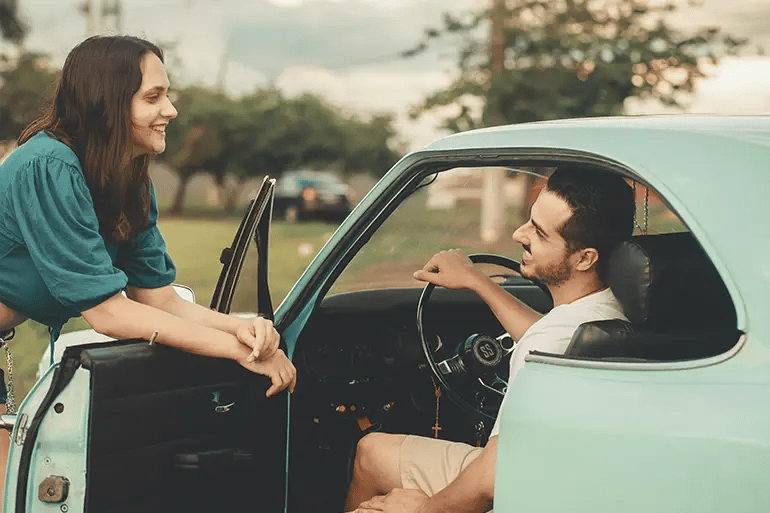Man in a blue car with the door open, smiling at a woman leaning on his door
