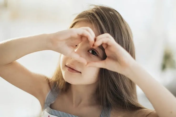A girl with brown hair and brown eyes making a heart symbol with her hands around her eye.