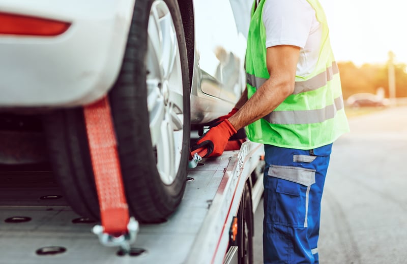 Tow truck operator wearing a high-visibility vest and protective gloves securing a car onto a recovery vehicle with safety straps during roadside assistance.