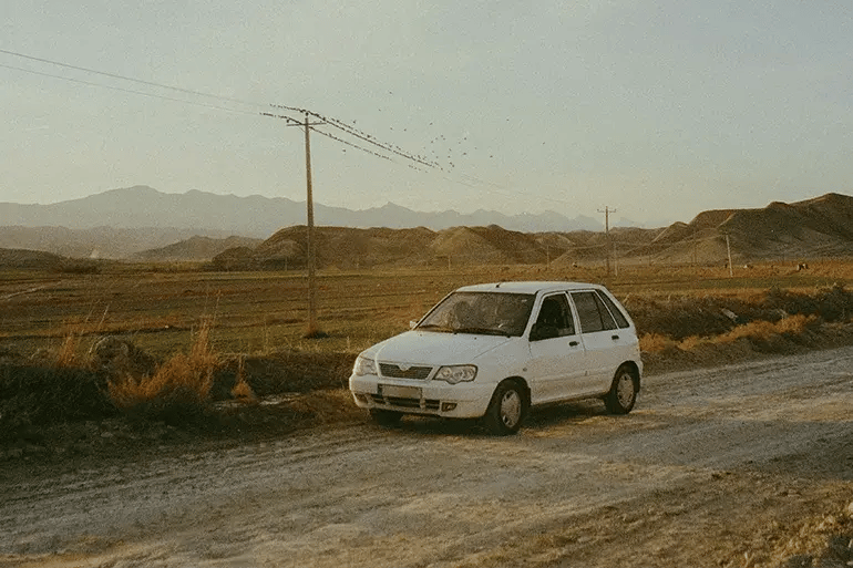 White compact car parked on a rural dirt road at sunset, surrounded by dry fields, low mountains and utility poles beneath a wide, pale blue sky.