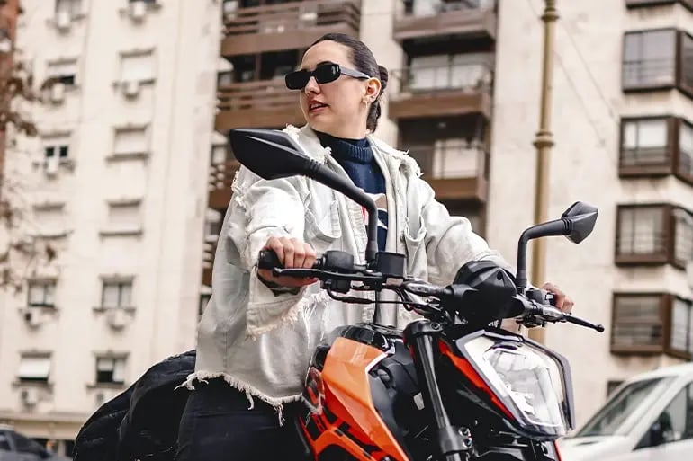 Image of a young girl with her hair tied back in a ponytail and wearing sunglasses, sitting on a motorbike. In the background, urban buildings can be seen.