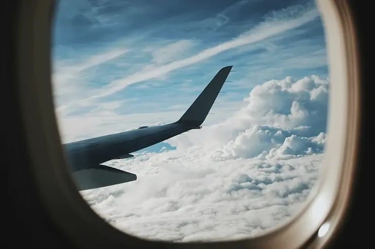 Aerial view from an airplane window in mid-flight, showing the wing of the aircraft above a sea of white clouds and a blue sky with contrails from other planes.