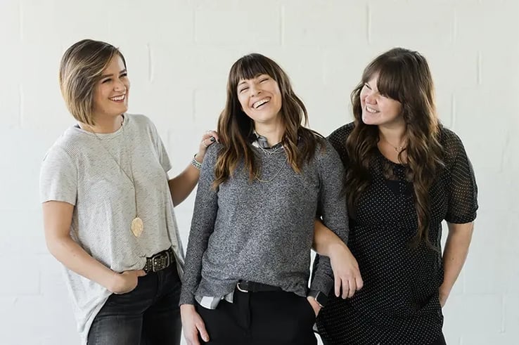 Three girls laugh as they look at each other, with a white background.