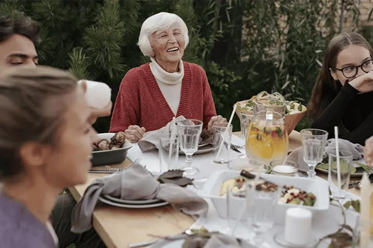 An elderly woman laughs enthusiastically as she shares a varied meal with her family, seated around a table. A scene full of affection, joy, and vitality for a lifetime.