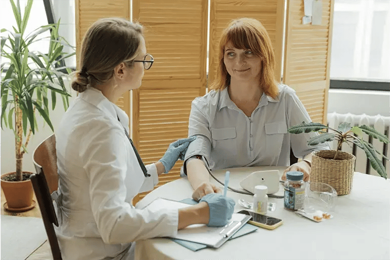 A red-haired girl is sitting at a table in a house with several floors. To her right, a doctor wearing a coat and gloves takes her blood pressure while writing down information. There are various medicines on the table.