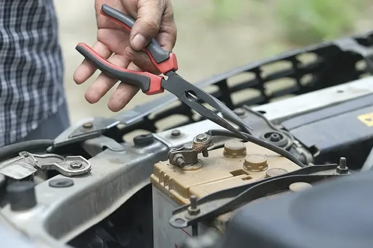 Person using pliers to manipulate a car battery, illustrating one of the potential problems of leaving a car unused, such as battery deterioration due to lack of use.