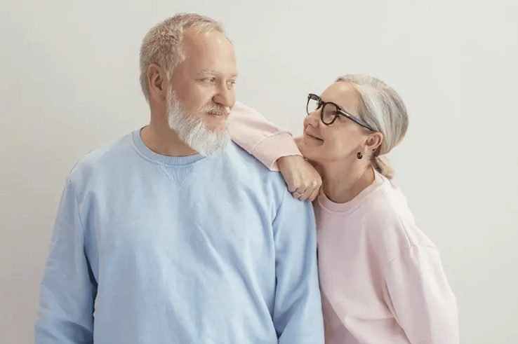 An elderly couple on a white background gaze tenderly at each other. The woman, wearing a pink jumper and black glasses, smiles at the man, who is wearing a blue jumper and has his elbow resting on her shoulder.