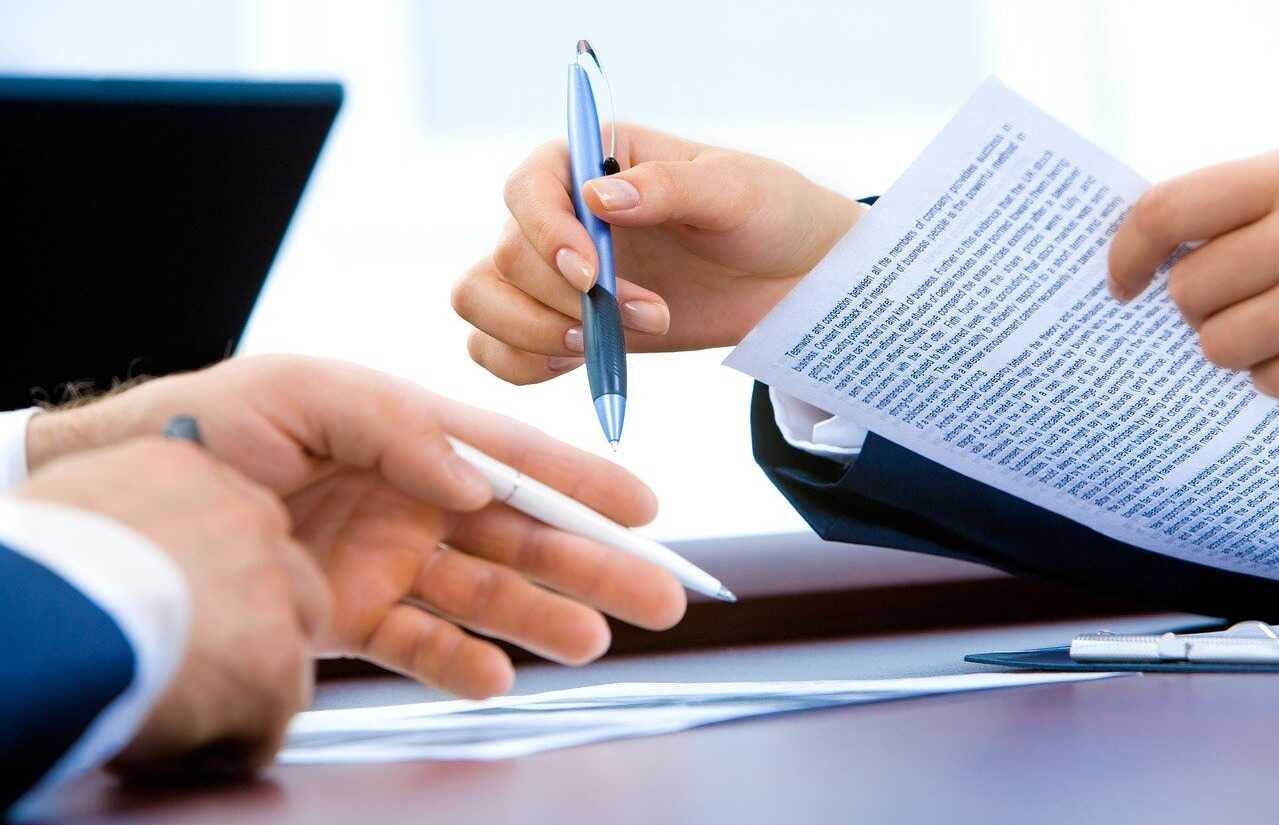 Image of two people's hands. One points to a document on the table with a pen, while the other holds a pen and a contract.