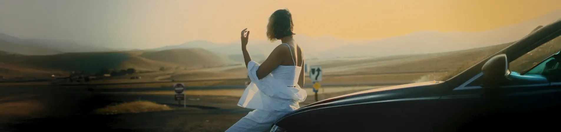 woman in a white dress sitting on the bonnet of a black car watching the sunset