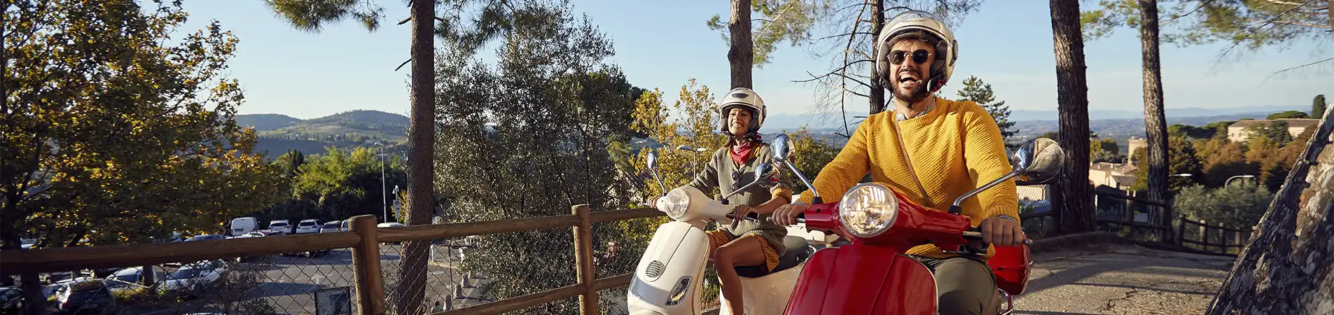 Image of a couple laughing as they ride a motorbike together at the start of a nature trail. Although they are already riding among trees, the car park near the start of the trail can still be seen in the background.
