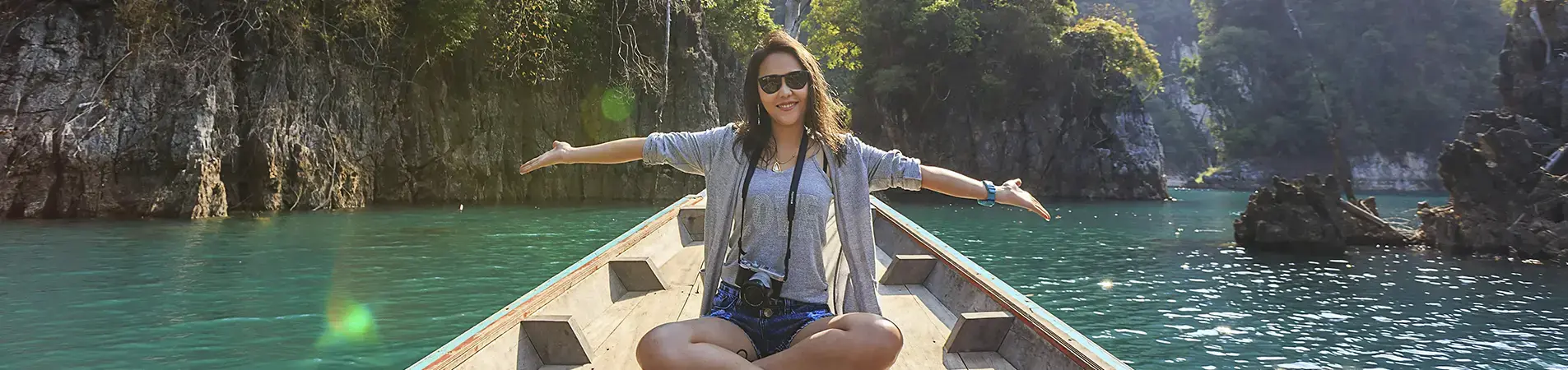 Young woman sitting at the bow of a wooden boat on a river or lake surrounded by rock formations and vegetation. She opens her arms in a gesture of enjoyment. She is wearing sunglasses and has a camera hanging around her neck.