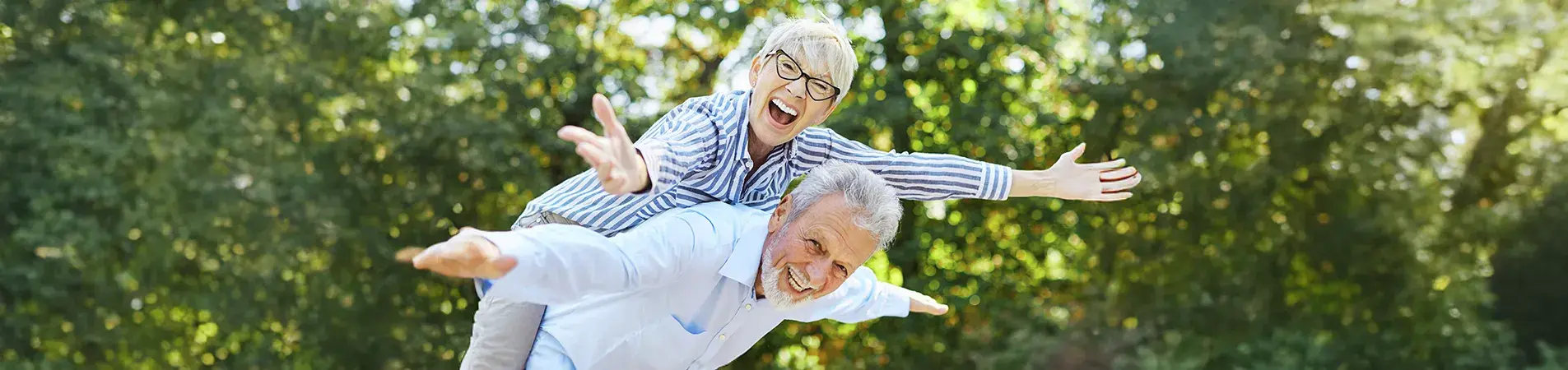 An elderly couple smiles at the camera with numerous trees in the background. The woman is standing on the man's back and both are spreading their arms as if they were aeroplane wings.